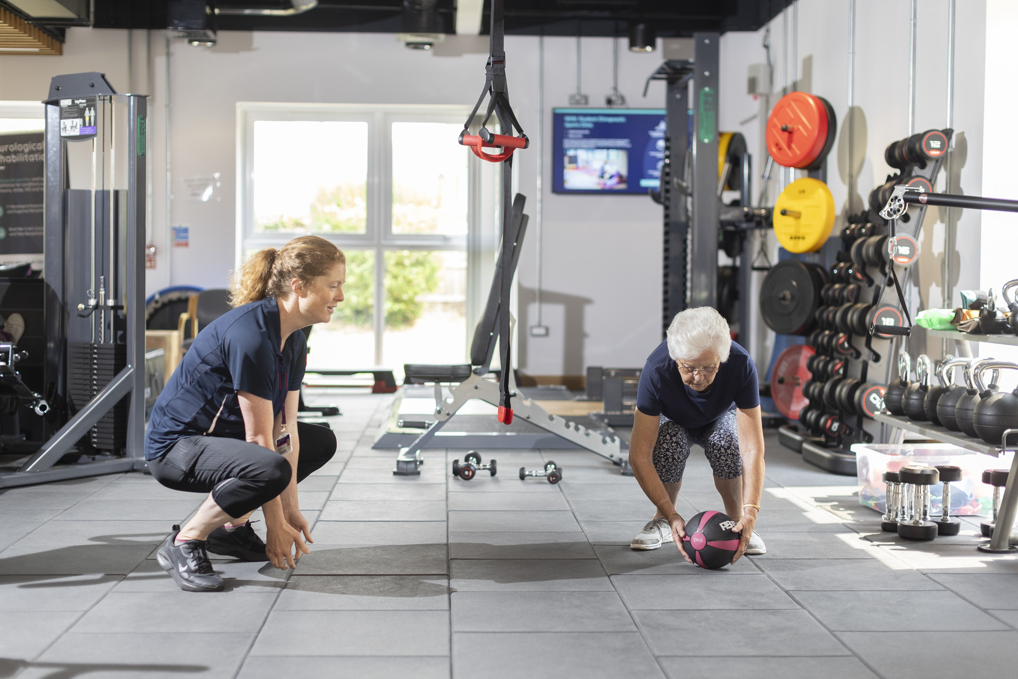 Female patient teaching an elderly patient squat technique involving a weighted ball