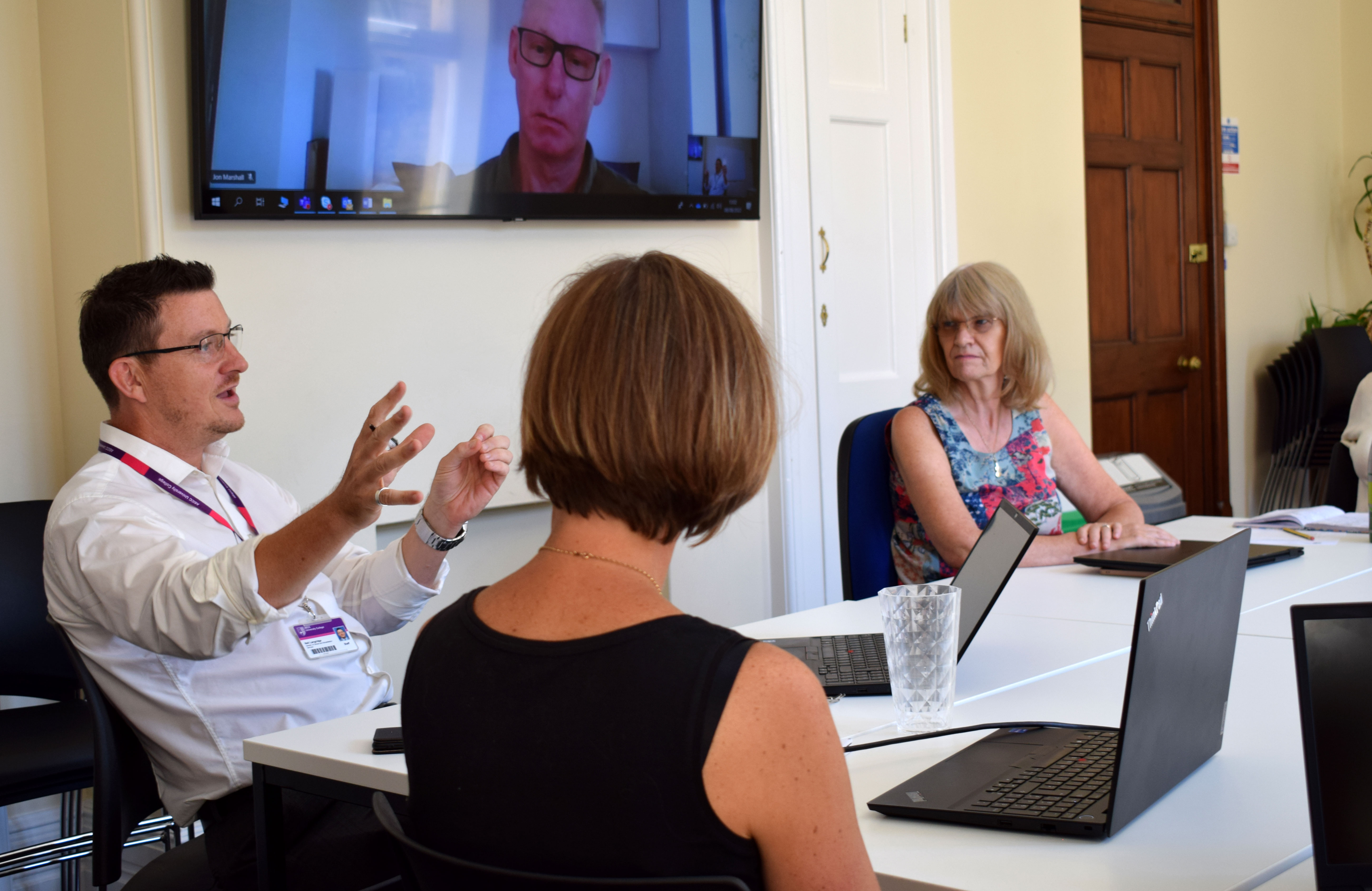 clinicians in a hybrid meeting with male colleague calling in remotely via a large screen and four colleagues in a meeting room