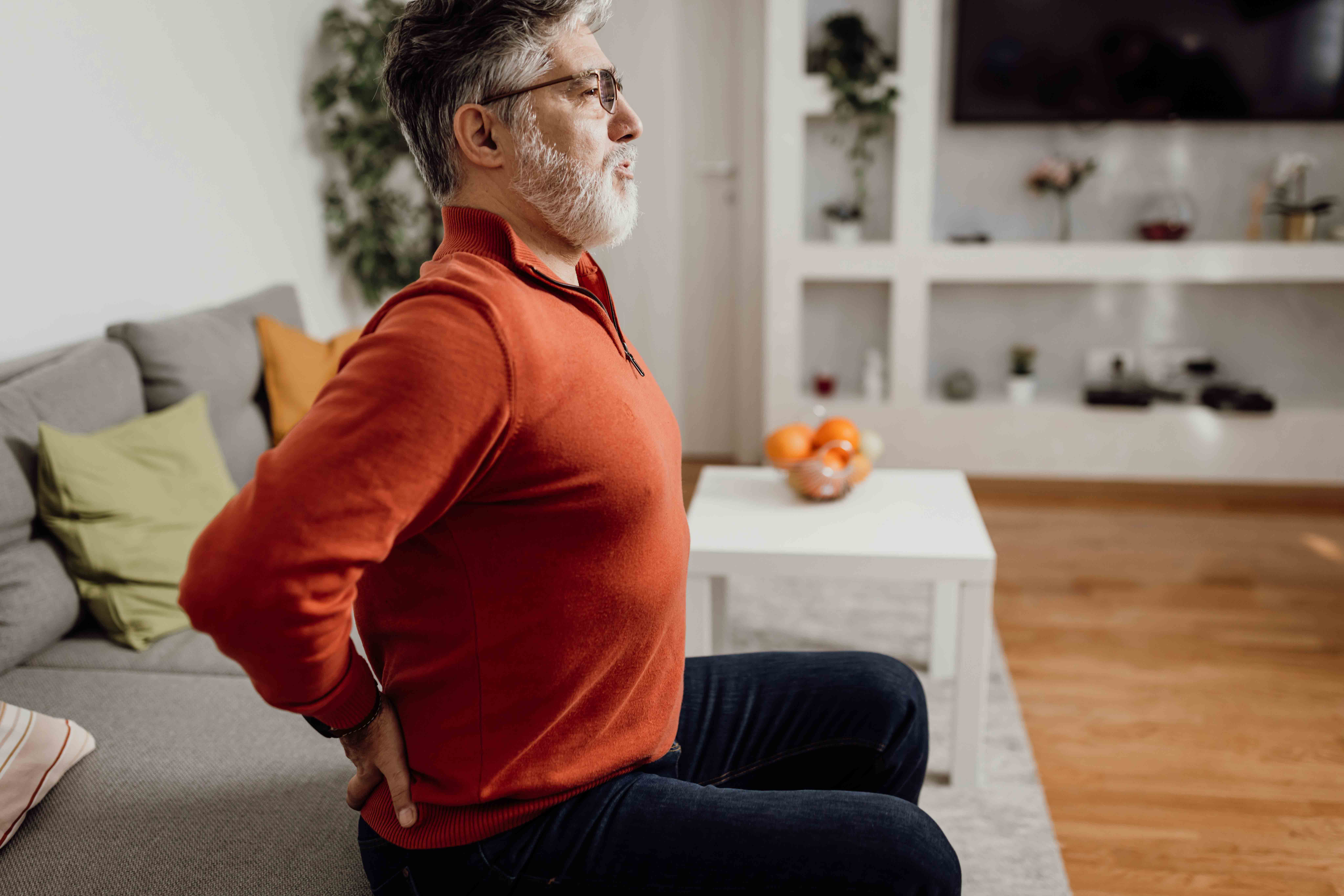 Side view worried mature man sitting at his living room couch holding hands on his painful lower back suggesting perhaps sciatica or other low back pain condition